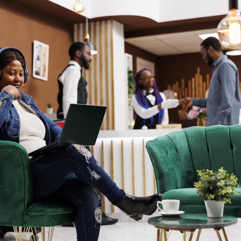 Young African American woman traveler sitting with laptop in hotel lobby using free wireless internet, watching movie online while waiting for check-in. Female freelancer working remotely from resort